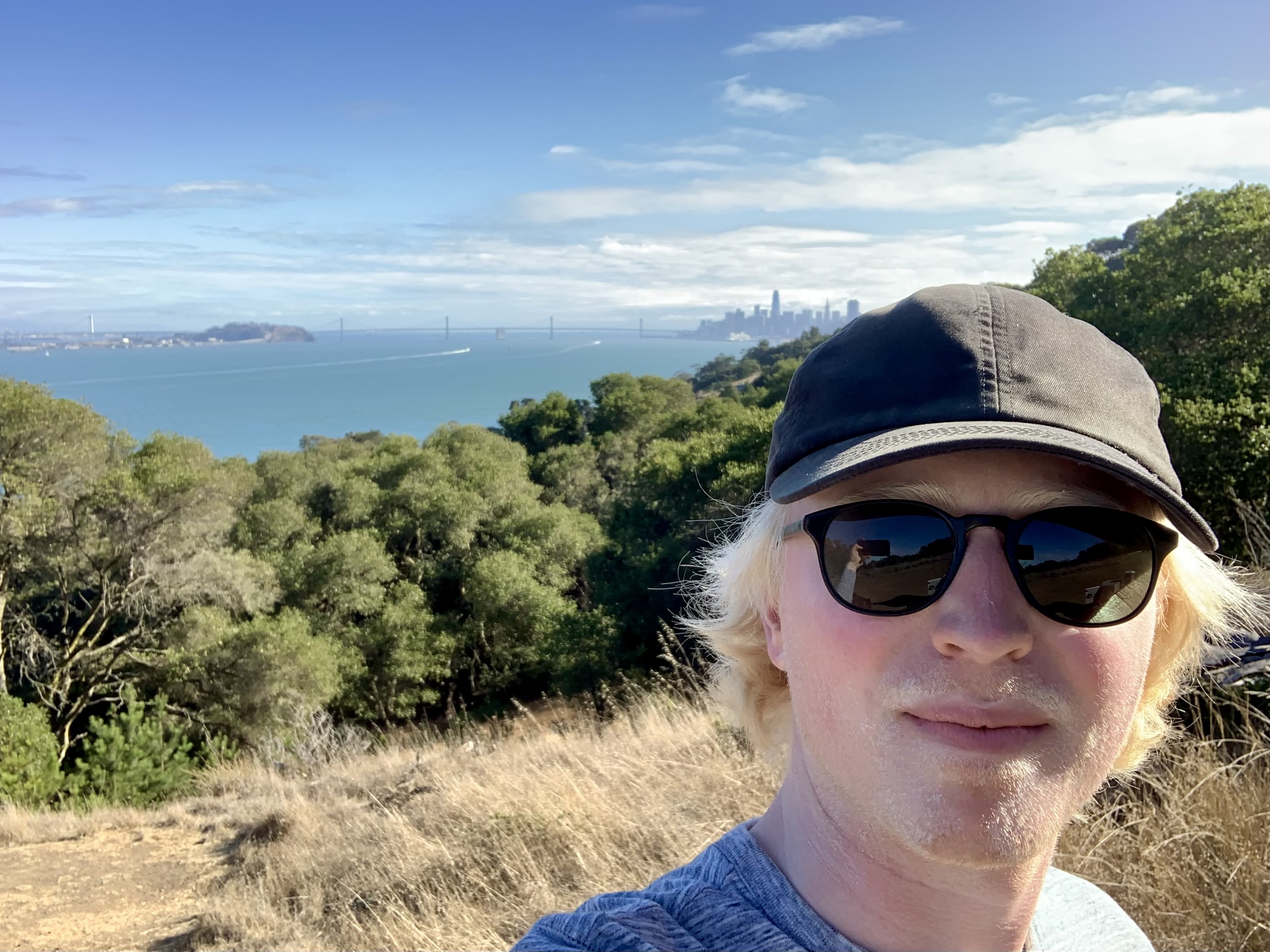 Face in foreground with baseball cap and sunglasses, Bay Bridge and SF cityscape in background. Taken from Angel Island